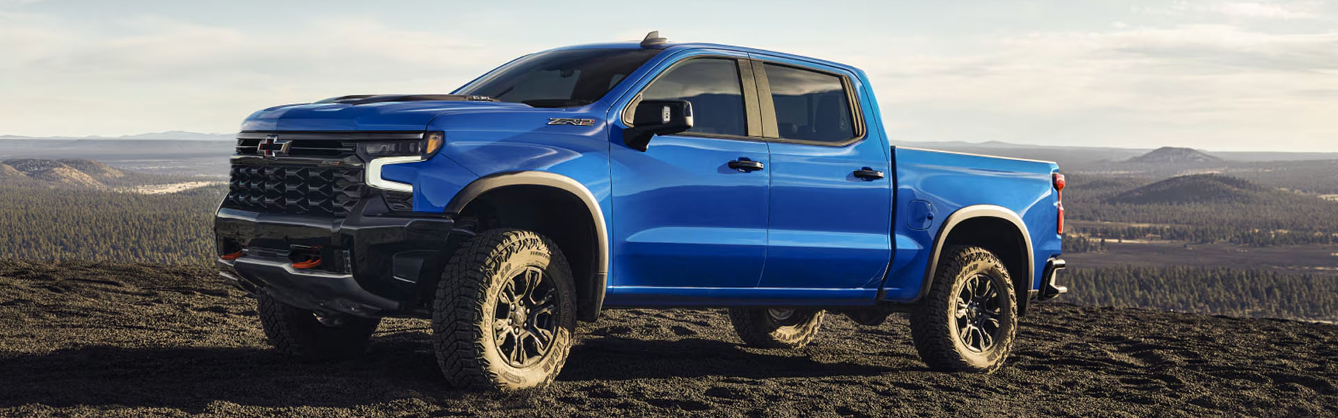 2026 Chevrolet Silverado in blue is parked on rugged off-road terrain against a mountainous landscape.