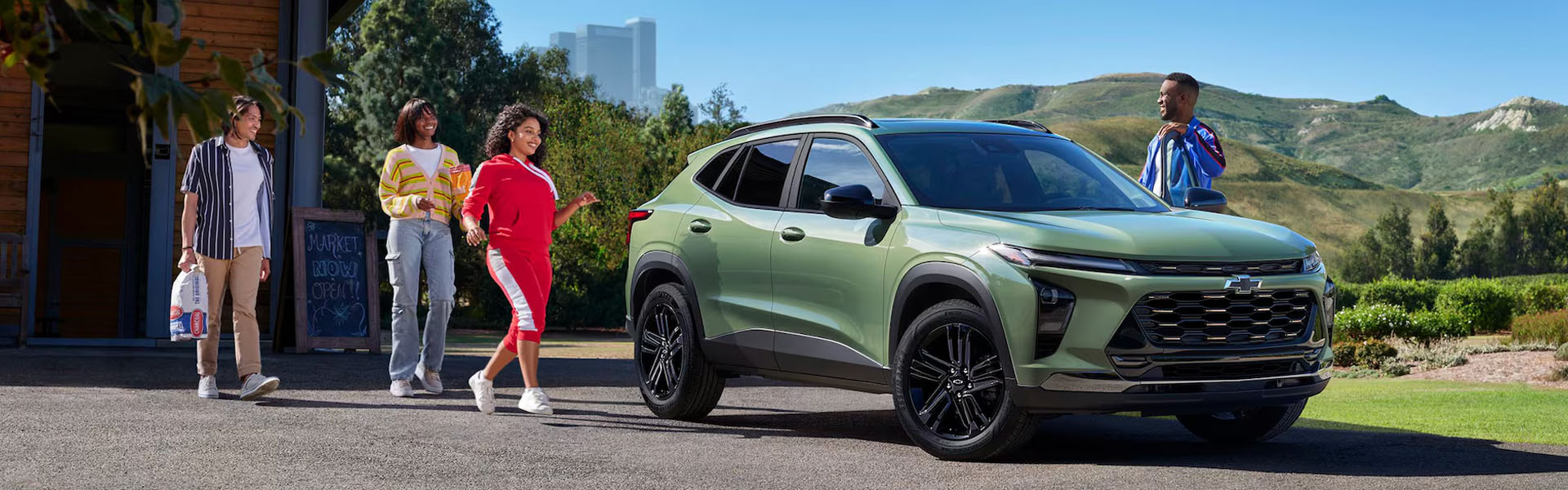 2026 Chevrolet Trax compact SUV in matte green parked outside a modern home, with a group of friends approaching—featuring sporty styling, black alloy wheels, spacious interior, and fuel-efficient design, ideal for urban driving and everyday versatility.