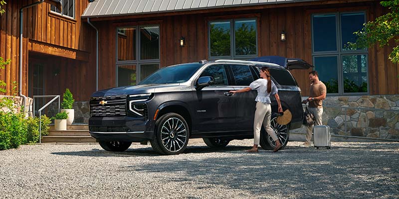 Couple loading luggage into a black Chevrolet Tahoe parked outside a modern wooden home, preparing for a trip on a sunny day.