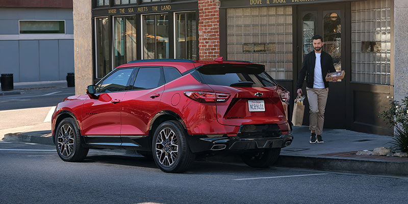 A red Chevy Blazer is parked on a city street near a brick building. A man with casual attire is holding takeout food, walking towards the vehicle.