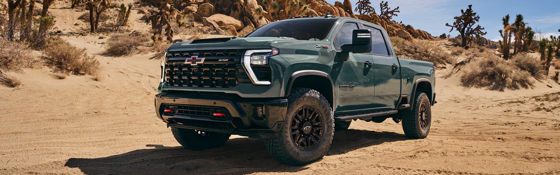 2026 Chevrolet Silverado HD heavy-duty pickup truck in green, shown in a desert landscape with rugged off-road tires and a bold front grille.