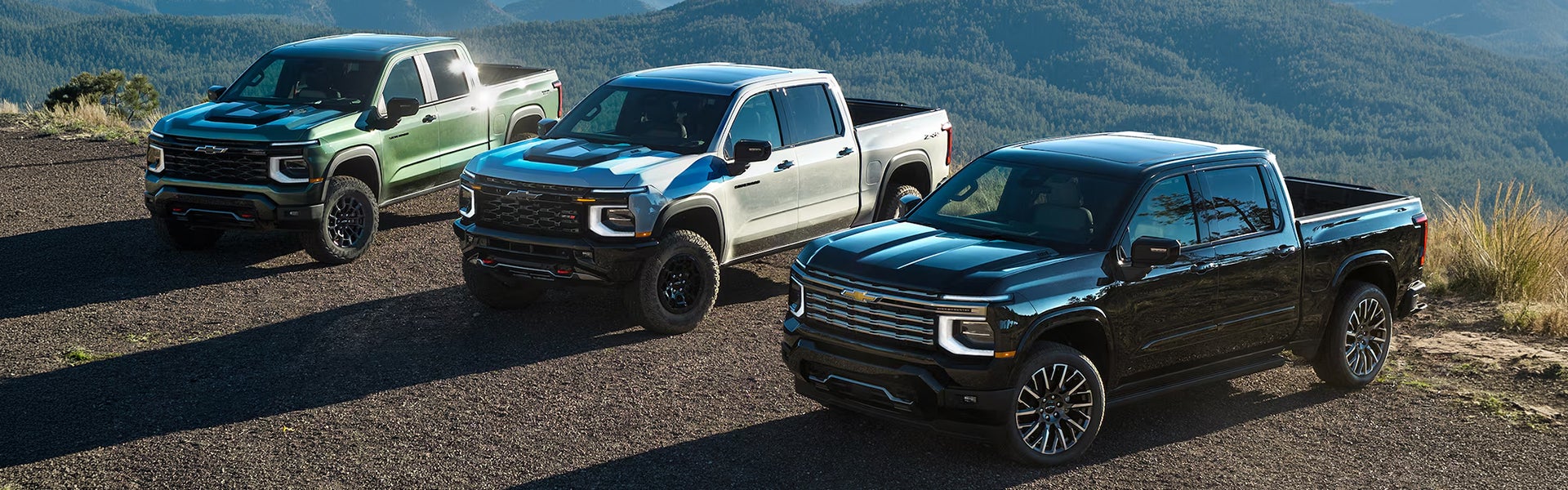 Three 2027 Chevrolet Silverado 1500 trucks in different colors parked on a scenic mountain landscape. 